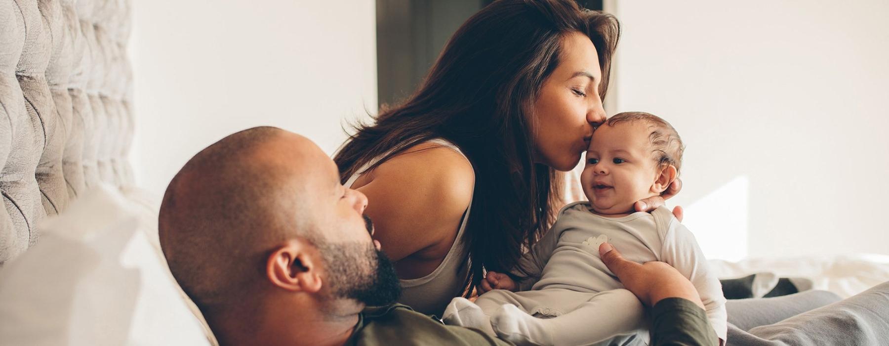 mother kisses her infant while father lies in bed and holds him
